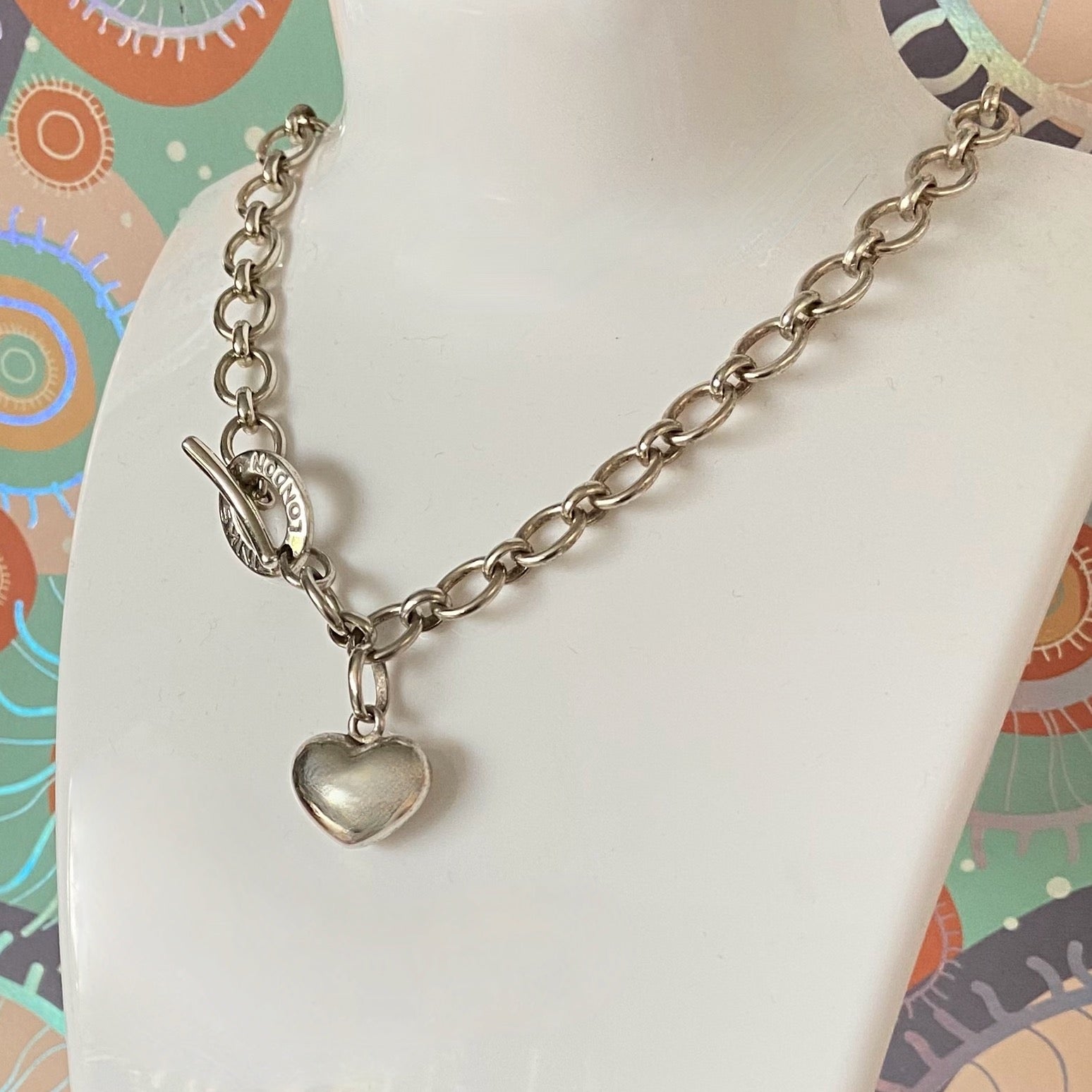 Silver chain bracelet with a heart charm on a mannequin arm against a colorful background