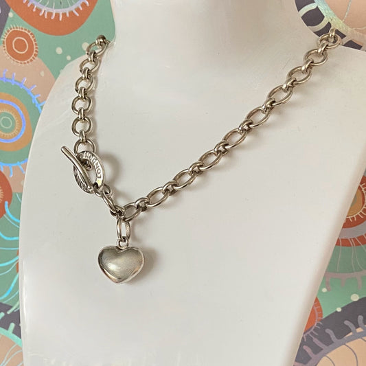 Silver chain bracelet with a heart charm on a mannequin arm against a colorful background